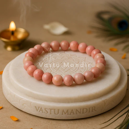 100% Natural Rhodochrosite beads with beautiful pink to rose-red coloration and natural banding patterns,  on a white stone platter with 'Vastu Mandir' engraving, lit diya in the background