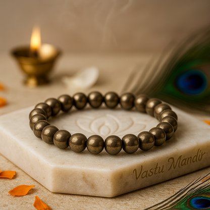natural pyrite stretch bracelet on a marble slab with 'Vastu Mandir' branding, candle, and peacock feather in the background.