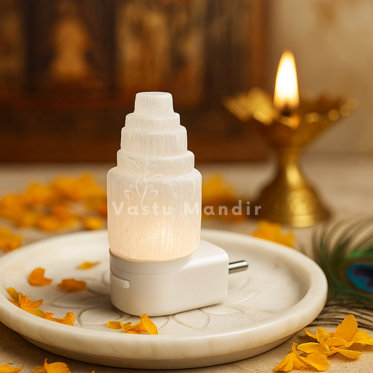 Selenite Tower Lamp Vastu Remedy in Bedroom for Negative Energy Removal Peaceful Sleep Aura Cleansing on a stone surface with 'Vastu Mandir' branding, surrounded by yellow flowers.