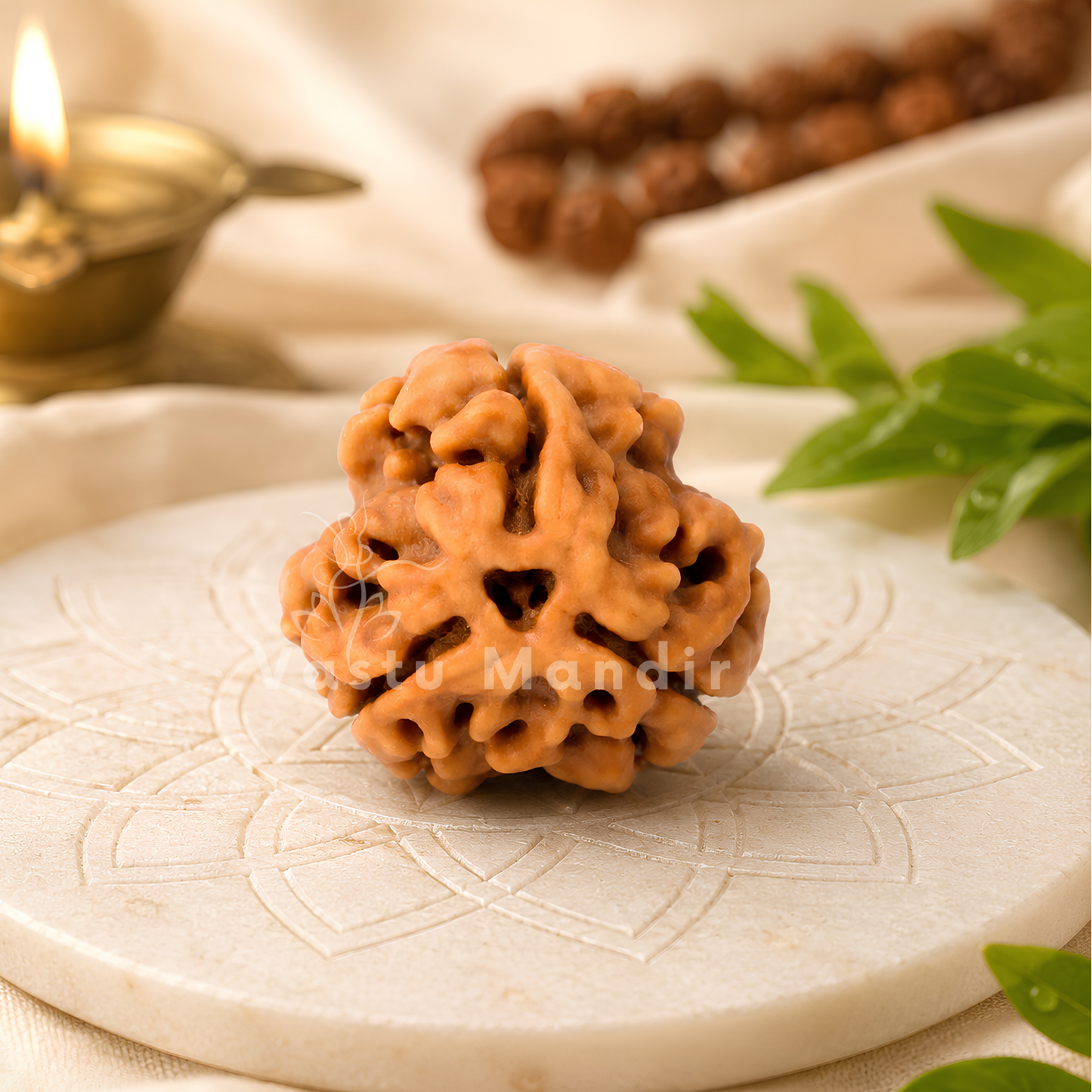 A carved wooden Rudraksha seed pendant sits on a round white marble tray, styled with a brass diya, green leaves, and prayer beads in the background.
