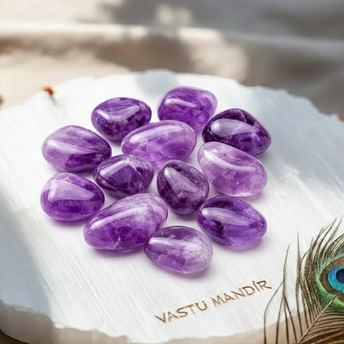 Amethyst stones on a white Selenite plate with 'Vastu Mandir' branding, accompanied by a peacock feather.
