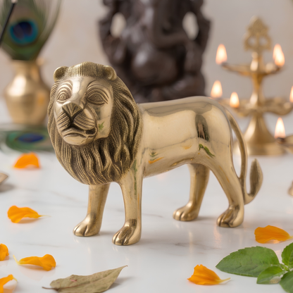 A polished brass lion figurine standing on a white marble surface, surrounded by scattered orange flower petals and green leaves. The background is softly blurred, showing decorative elements like a brass diya holder and a peacock feather.