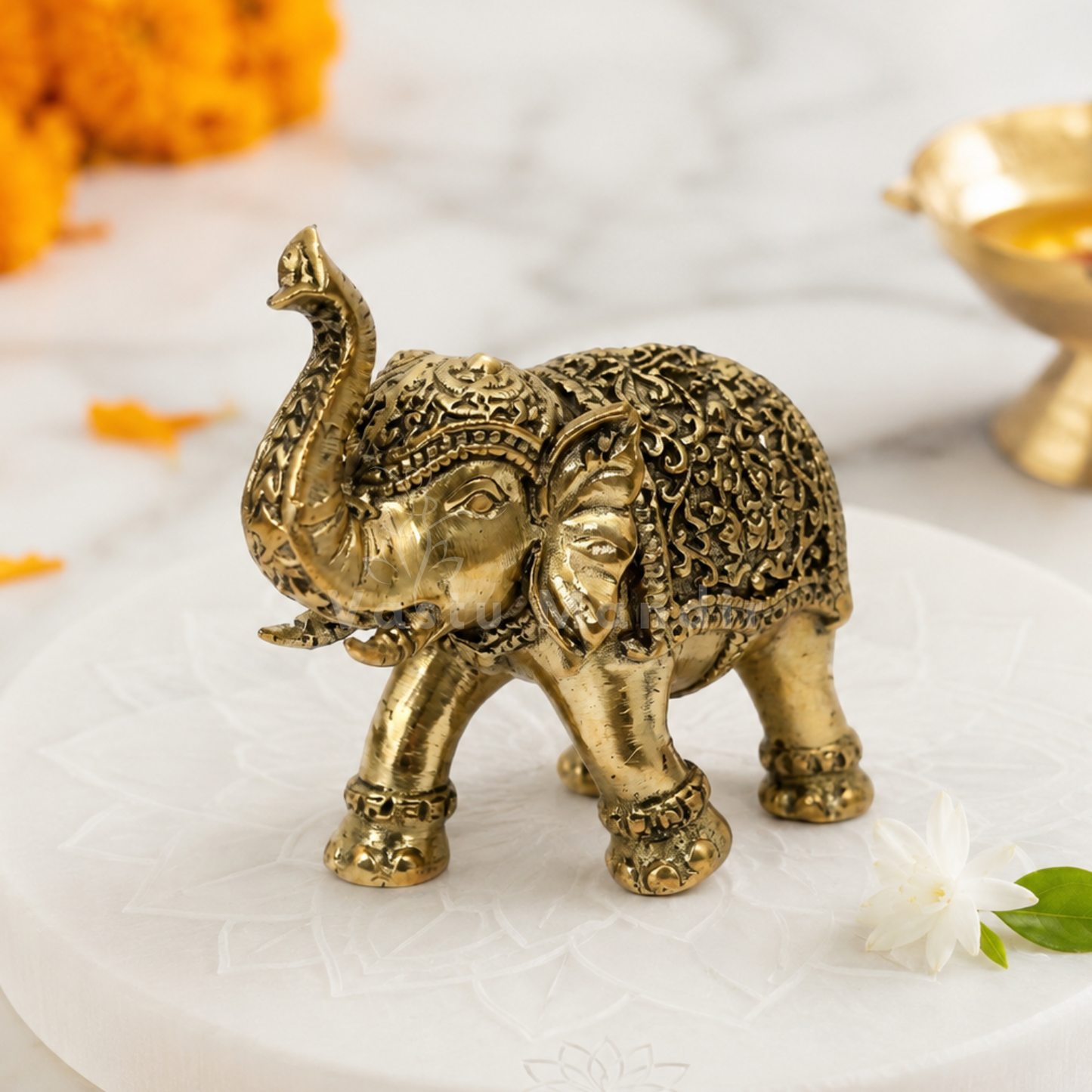 Decorative brass elephant figurine on a white surface with flowers and a bowl in the background.