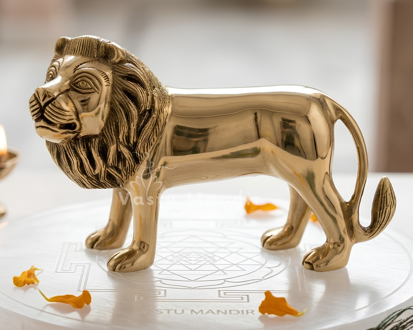 A polished brass lion figurine with a golden finish, standing on a white marble plinth decorated with small orange flower petals. A small lit brass diya candle is visible in the soft-focus background on the left.