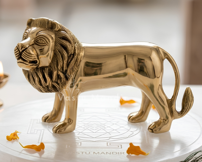 A polished brass lion figurine with a golden finish, standing on a white marble plinth decorated with small orange flower petals. A small lit brass diya candle is visible in the soft-focus background on the left.