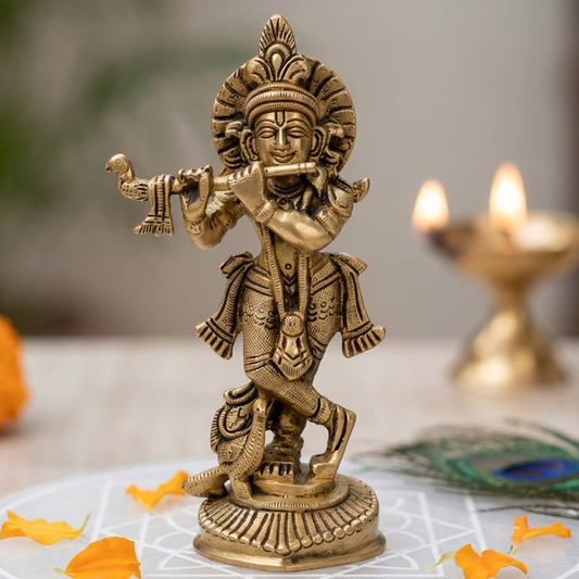 A brass Krishna Ji idol playing a flute on a white surface with candles and marigold flowers in the background.