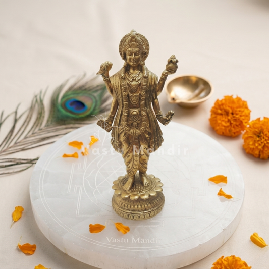 Brass statue of a Dhanvantri on a white platform with marigold flowers and a peacock feather.