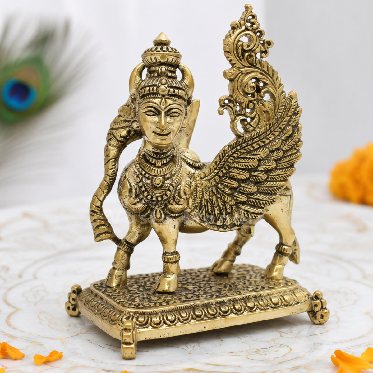 Flying Kamdhenu cow brass idol on a textured surface with peacock feathers in the background