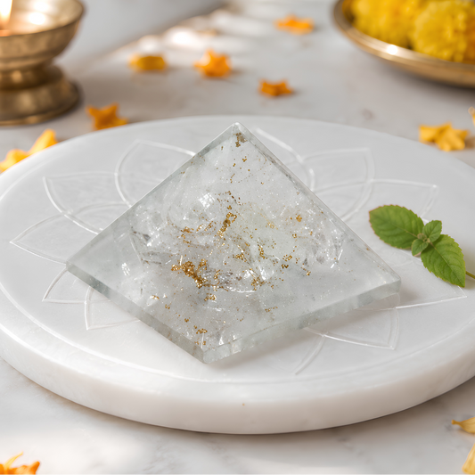 A clear Selenite crystal pyramid 1 inch with embedded gold flakes sits on a white marble tray, styled with a green mint leaf, scattered orange flower petals, a lit brass candle holder, and yellow decorative balls in the background.